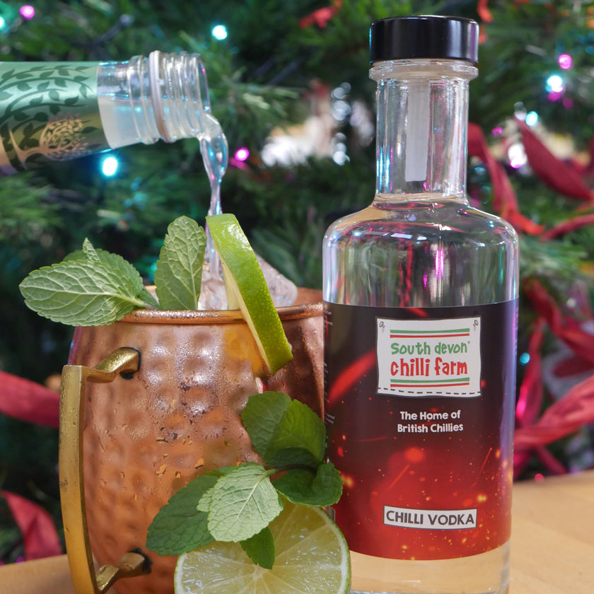 Bottle of Gin with lime slices and mint leaves on a wooden surface with a Christmas tree in the background.